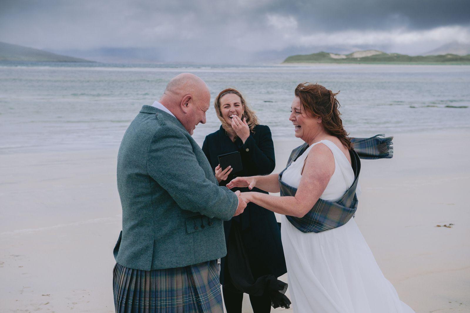 Bride and groom holding hands with their celebrant, all laughing, with Seilebost beach behind