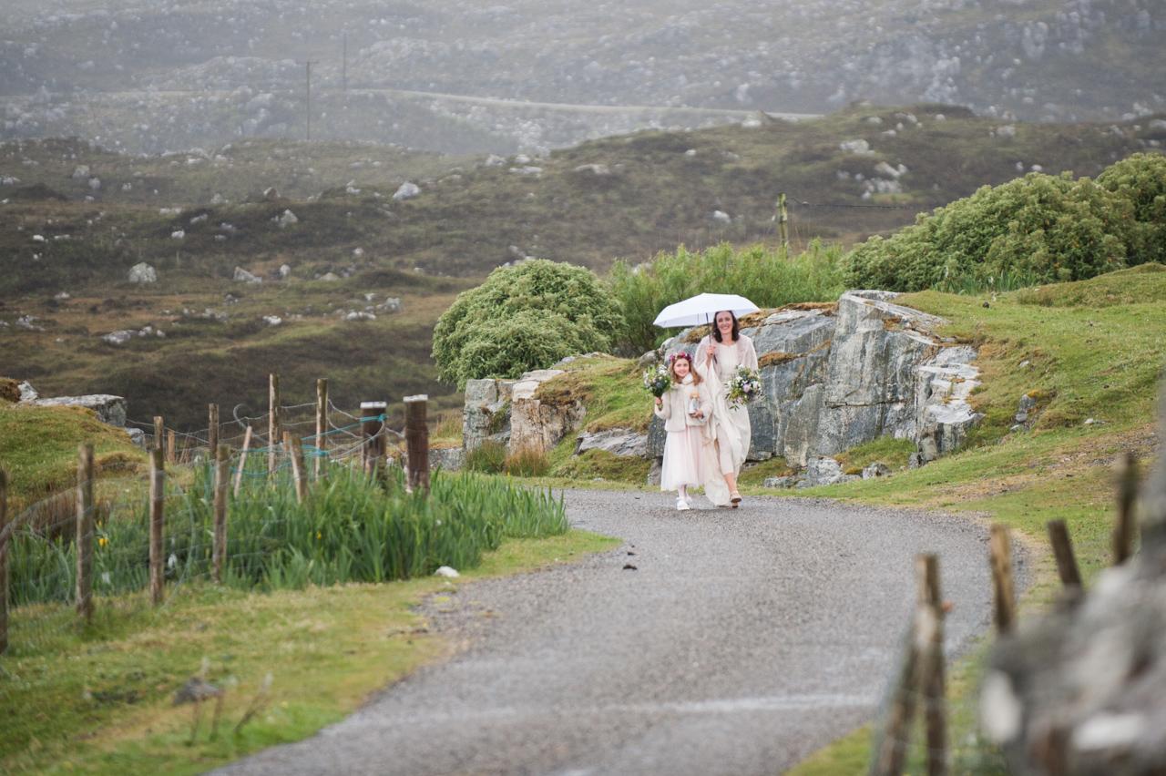 Emma walks along a lane on the Isle of Harris, with a white umbrella protecting her wedding gown from the rain. 