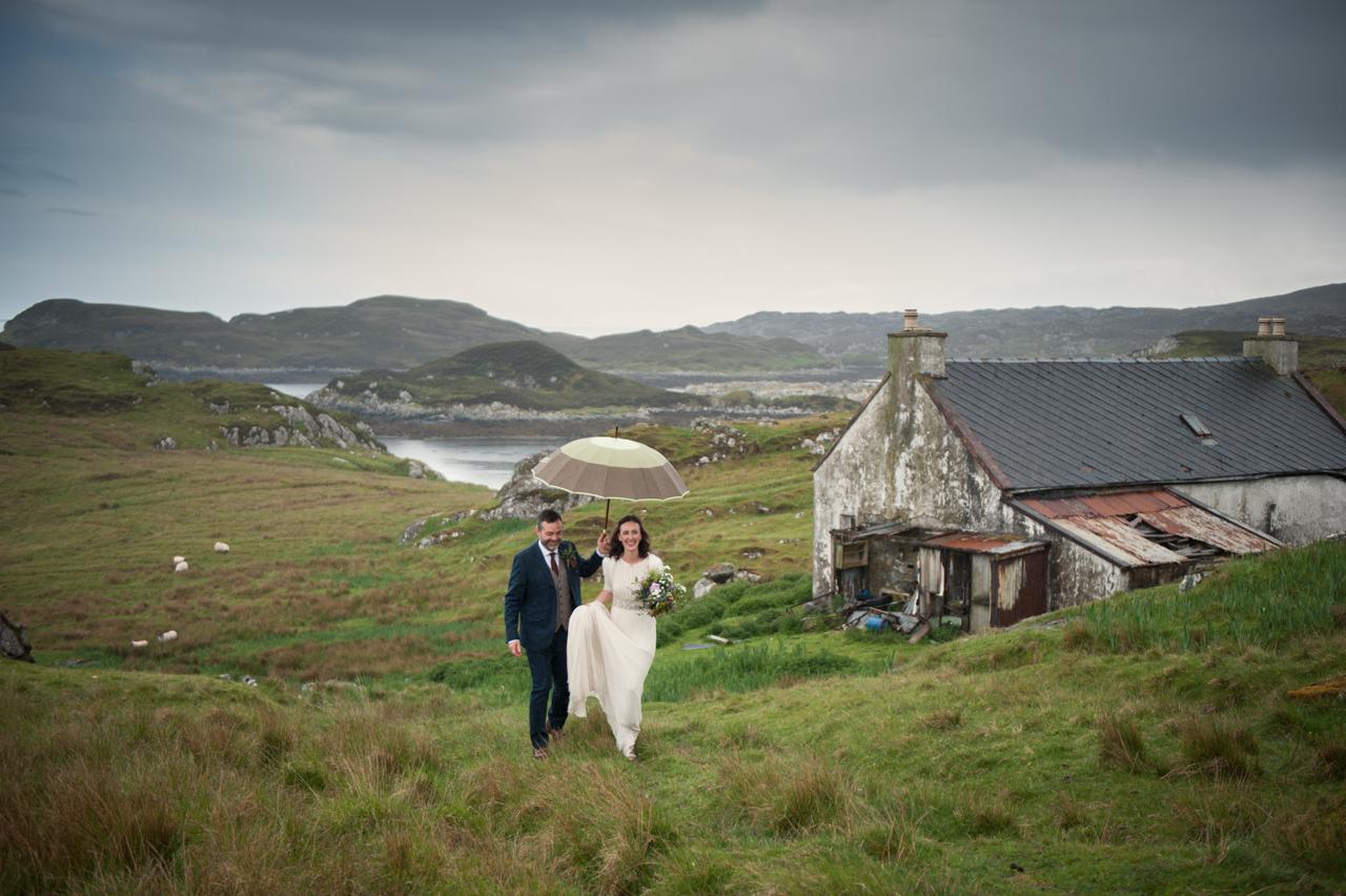 Emma and Jamie walk across a green field with sheep in the background. Jamie is holding am umbrella to shield Emma from the rain and she's carrying a bridal bouquet. 