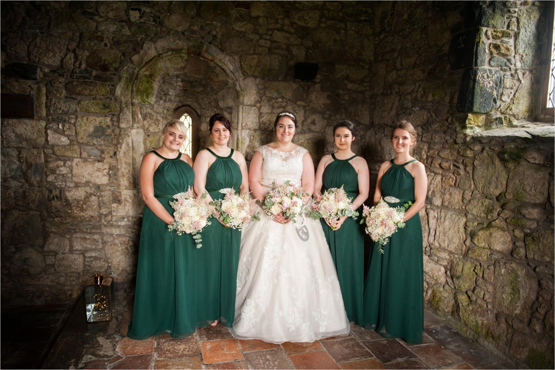 The bride and bridesmaids wearing emerald green pose for a formal portrait against a stole wall in the historic St Clements Church, Rodel, Isle of Harris. 