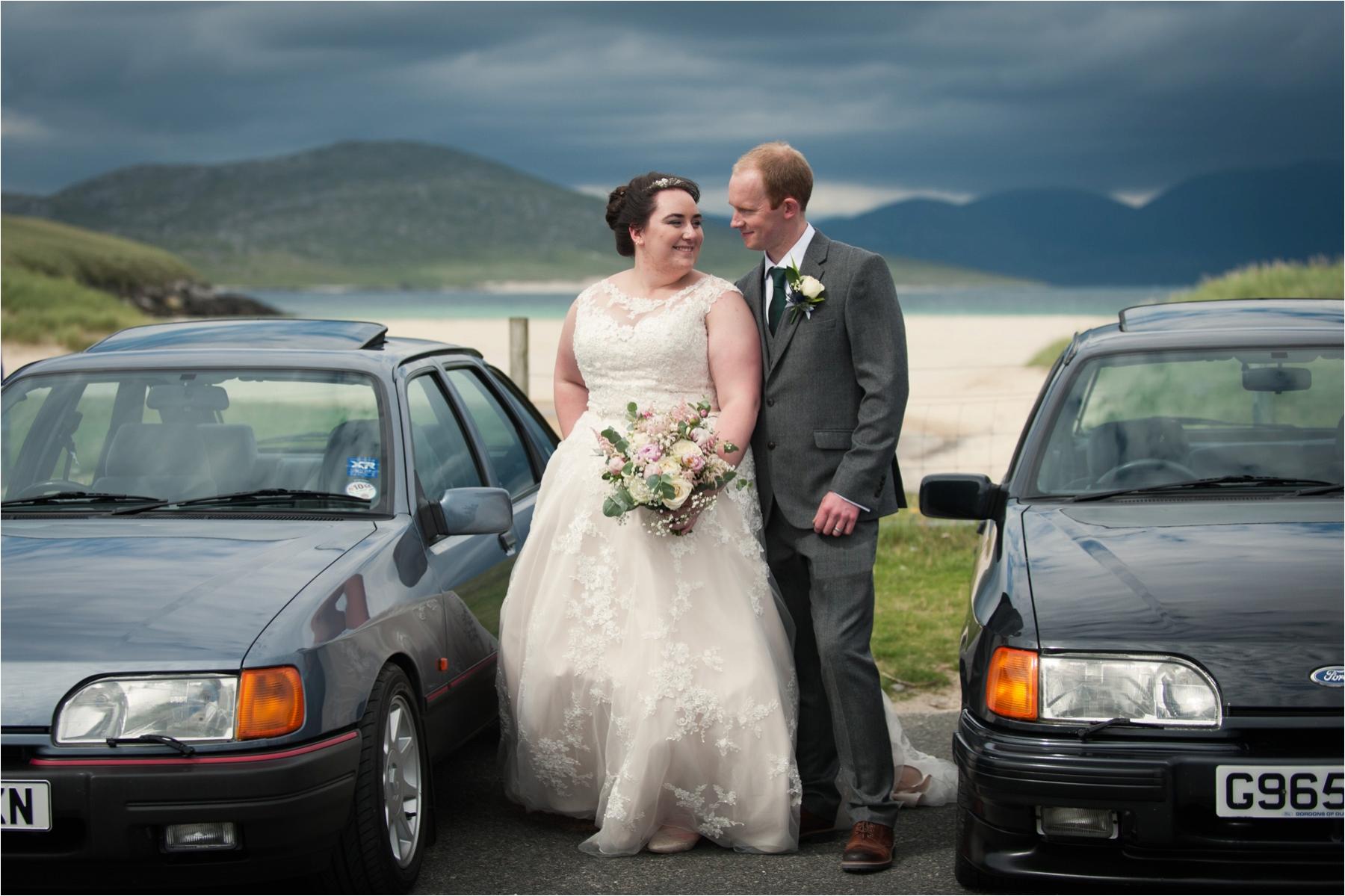 Bride and groom Lauren and Michael pose between two classic Ford cars near Horgabost Beach in Scotland. 