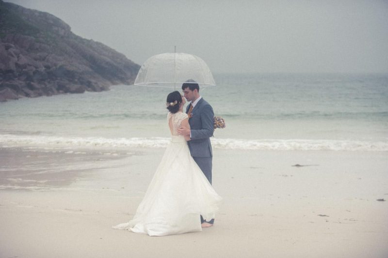 A bride in a full-length white dress and a groom snuggle under an umbrella on the Isle of Mull on their wedding day.