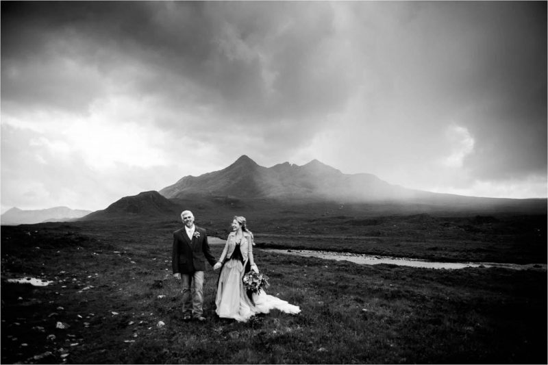 A bride and groom stand outdoors after their Skye wedding in this black and white photo showing a rain downpour in the background. Photographer: Margaret Soraya