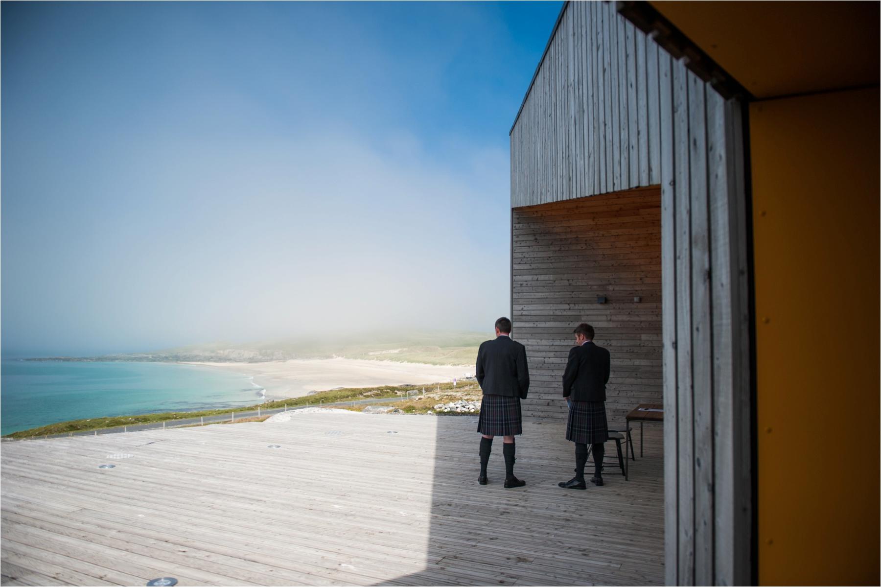 The groom and his best man wait at Talla Na Mara on the Isle of Harris before his wedding ceremony. They're overlooking a blue bay and there are blue skies. 
