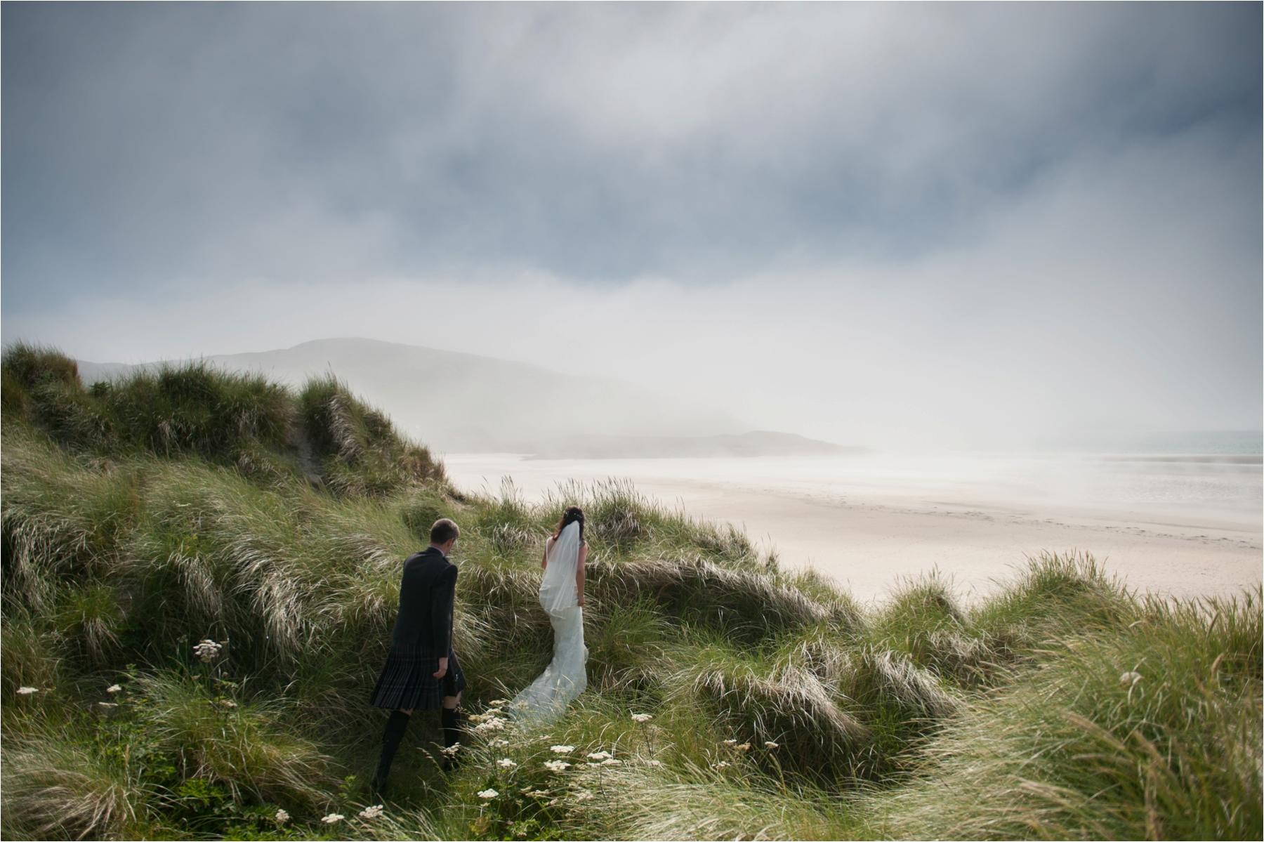 A bride and groom walk through the sand dunes of Luskentyre Beach in the Scottish Highlands. 