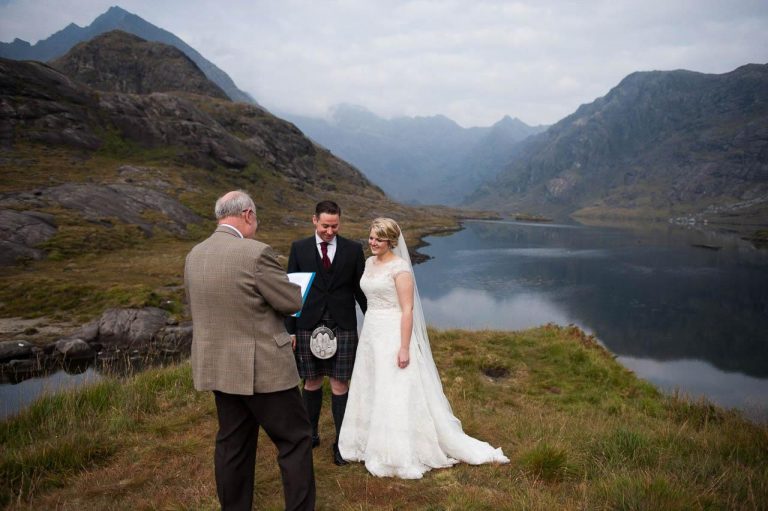 A civil wedding celebrant conducts an outdoor marriage ceremony on Loch Curuisk on the Isle of Skye. Behind the bride and groom is Loch Coruisk and misty mountains.