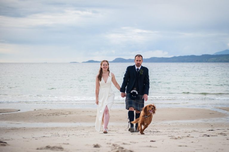 A bride and groom hold hands on a beach in the Scottish Highlands. Photographer:Margaret Soraya
