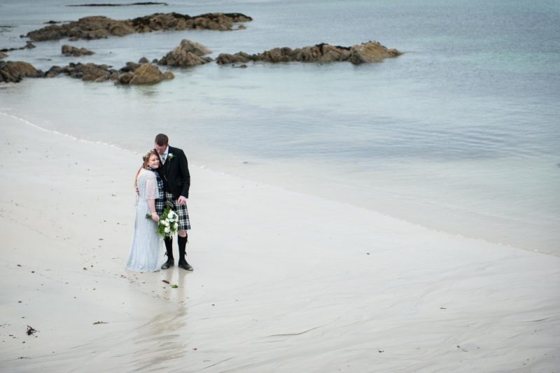 A bride in a full-length lavender dress and a groom in a black and white tartan kilt on a beach on Tiree for their wedding.
