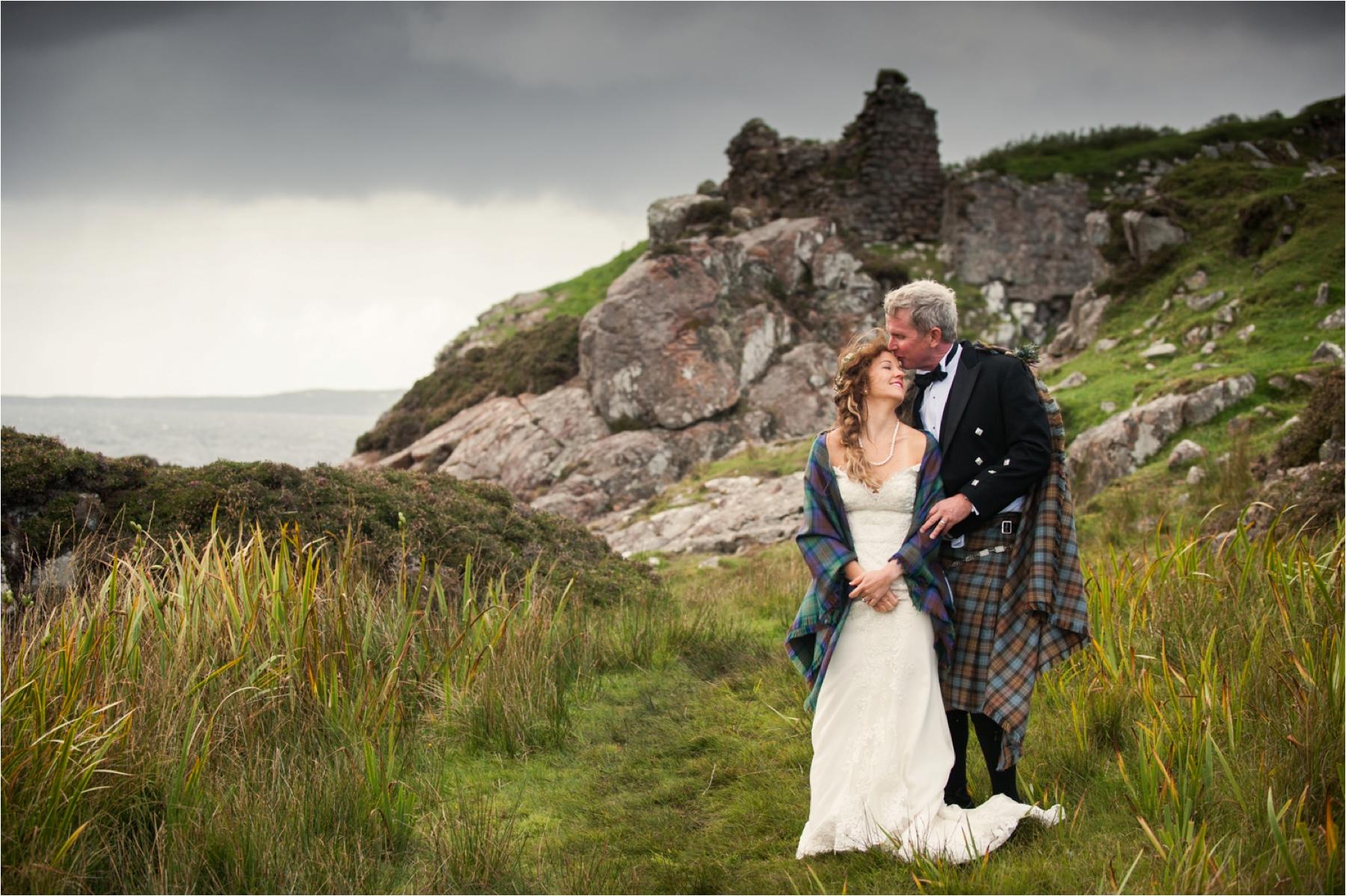 A Scottish elopement at Dunscaith Castle on the Isle of Skye with the groom kissing the head of the bride. She is wearing a tartan wrap to stay warm on the showery day. 